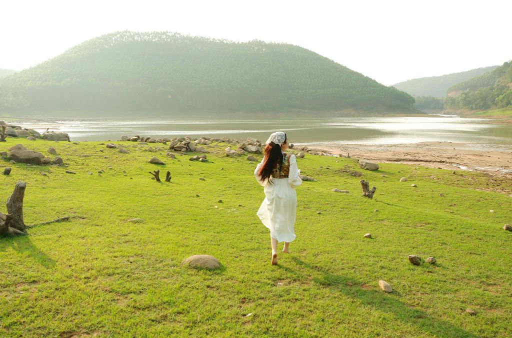 Người mặc váy trắng đứng trên bãi cỏ ven hồ, phía xa là những dãy đồi