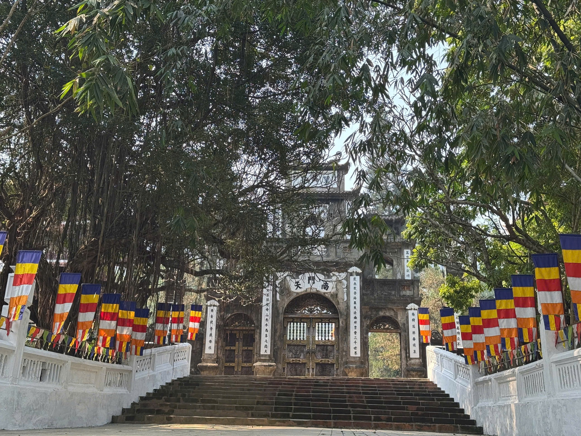 Cổng tam quan cổ kính tại Chùa Hương Hà Nội với bậc thang đá, hàng cây xanh và cờ Phật giáo hai bên lối vào.